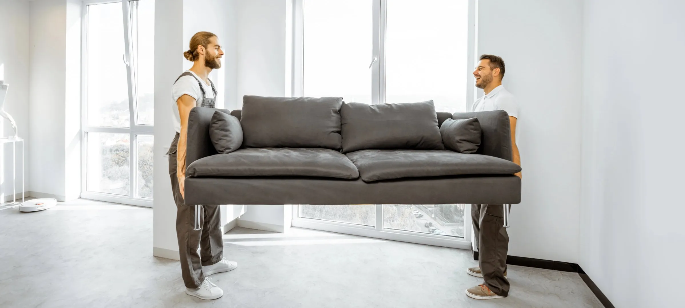 Two movers carrying a grey sofa into a bright, empty apartment