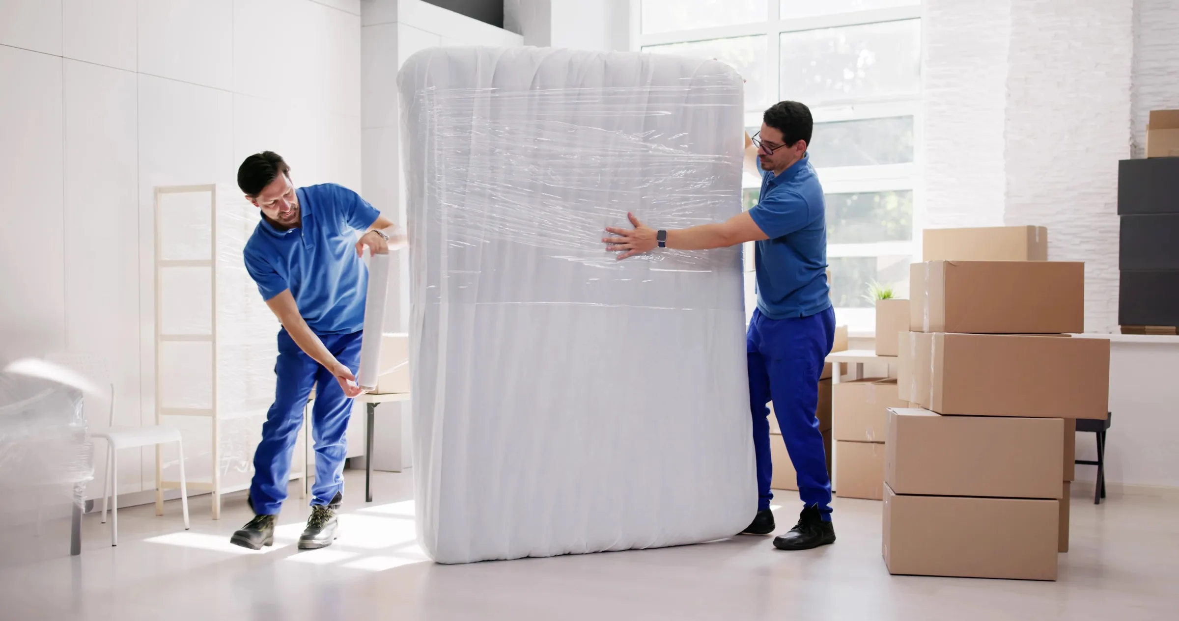 Two movers wrapping a mattress in plastic film before a move